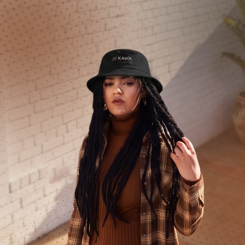 A woman with long dreadlocks wearing a black KAWA branded bucket hat, brown turtleneck, and plaid shirt against a white brick wall.